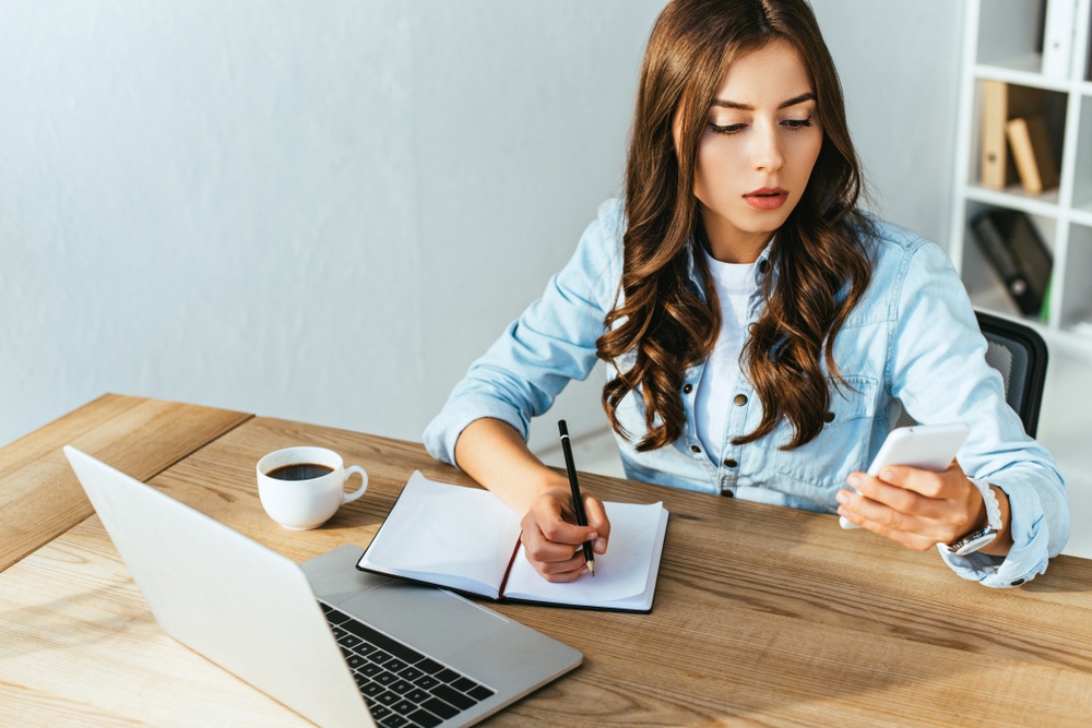 girl looking at her phone while shes at her desk writing in a notebook. computer open and coffee at the table