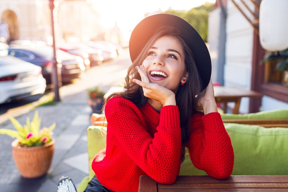 brunette woman with black fedora hat sitting at table pondering and smiling